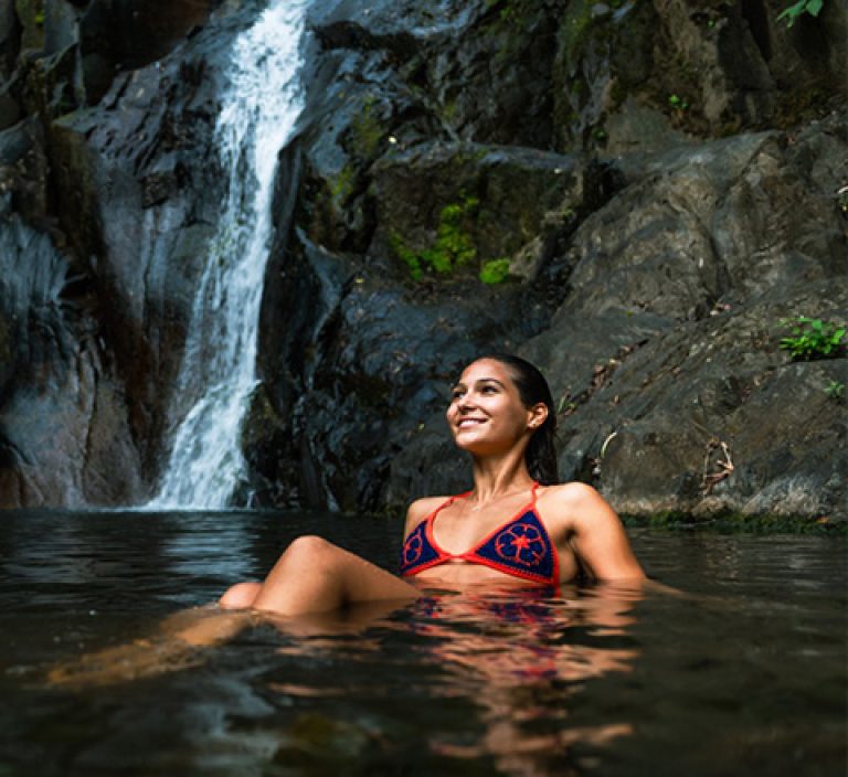 a woman smiles while lounging in water near a waterfall